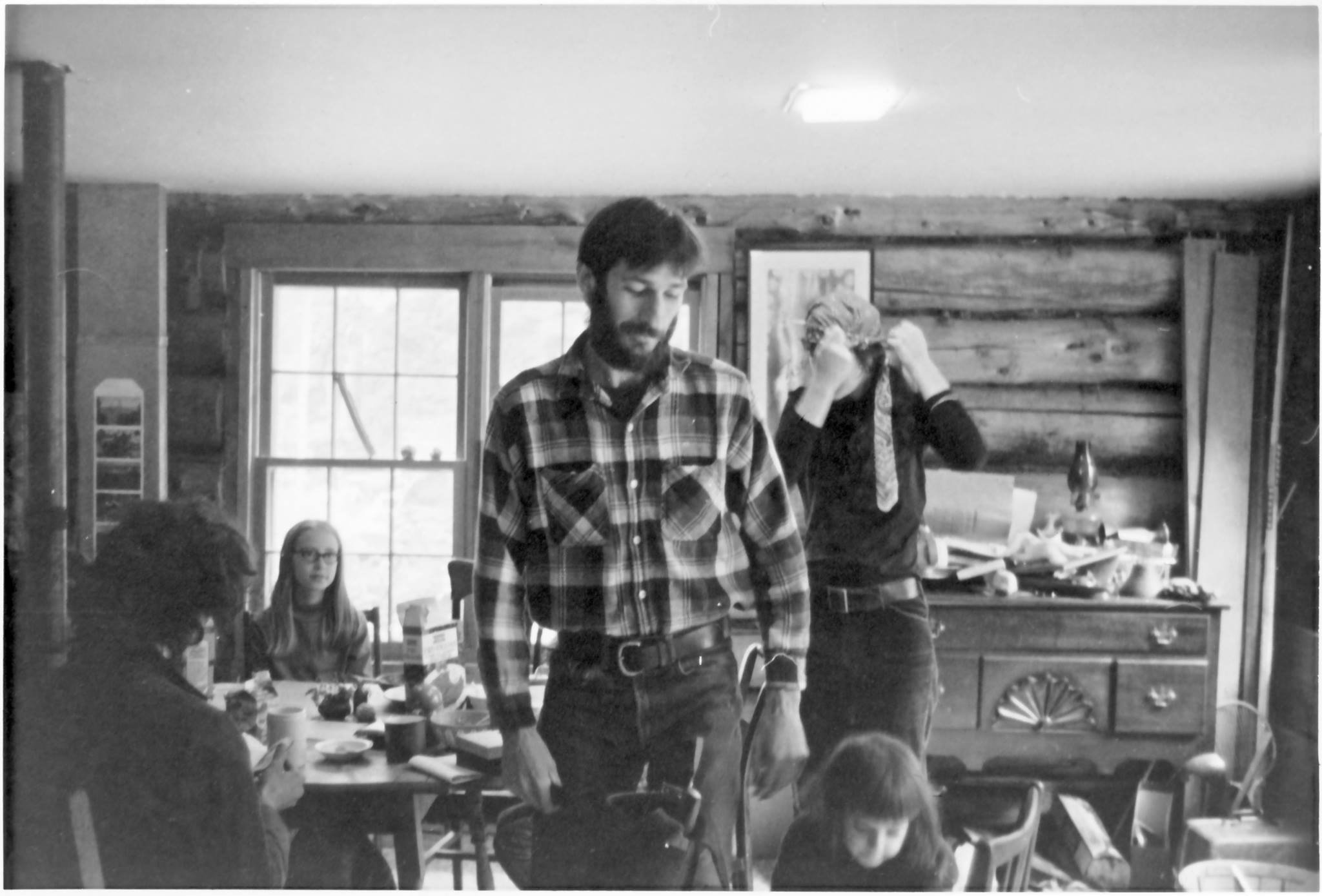 Black and white image of a lively gathering in a rustic cabin, featuring a man in a plaid shirt standing, a woman adjusting a scarf, and two children at a table with food and drinks. The scene captures a warm, familial atmosphere with wooden beams and a cluttered background, highlighting a cozy domestic setting.
