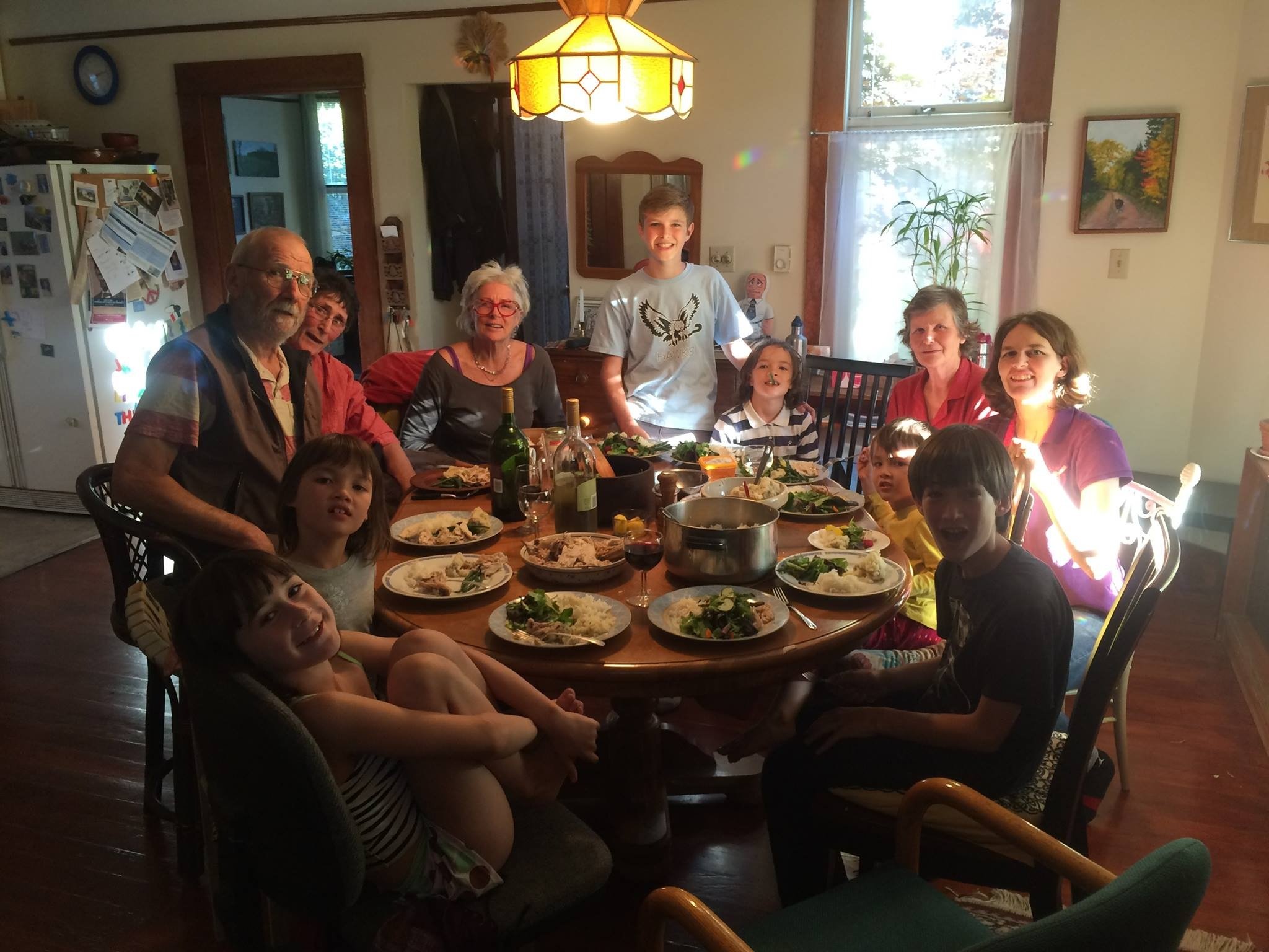 A lively family dinner scene featuring multiple generations gathered around a wooden table filled with plates of food, including salads and main dishes. The warm atmosphere is enhanced by natural lighting, showcasing the joy of togetherness in a cozy dining room setting.