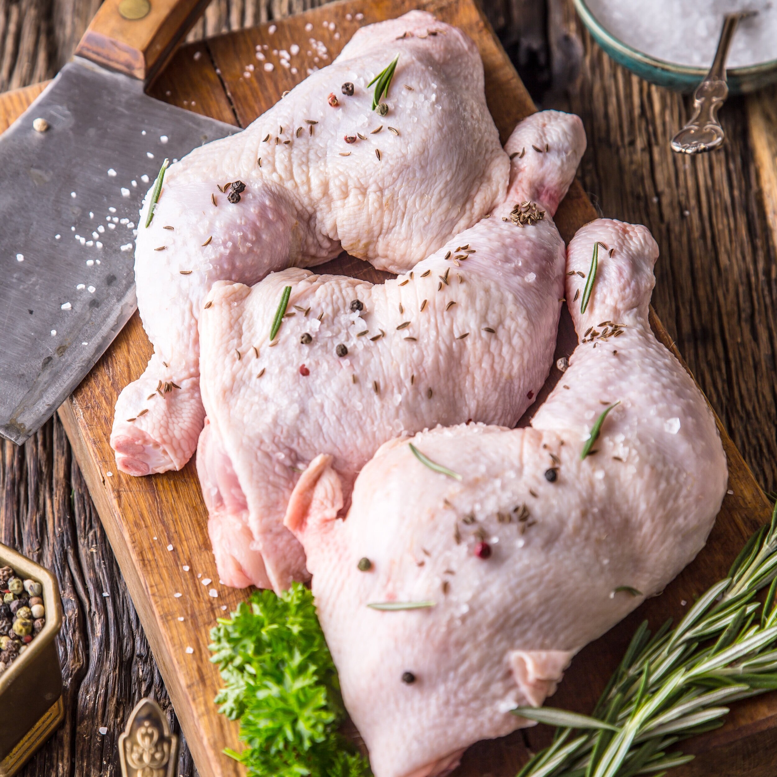 Raw chicken thighs seasoned with herbs and spices on a wooden cutting board, accompanied by a cleaver and fresh parsley.