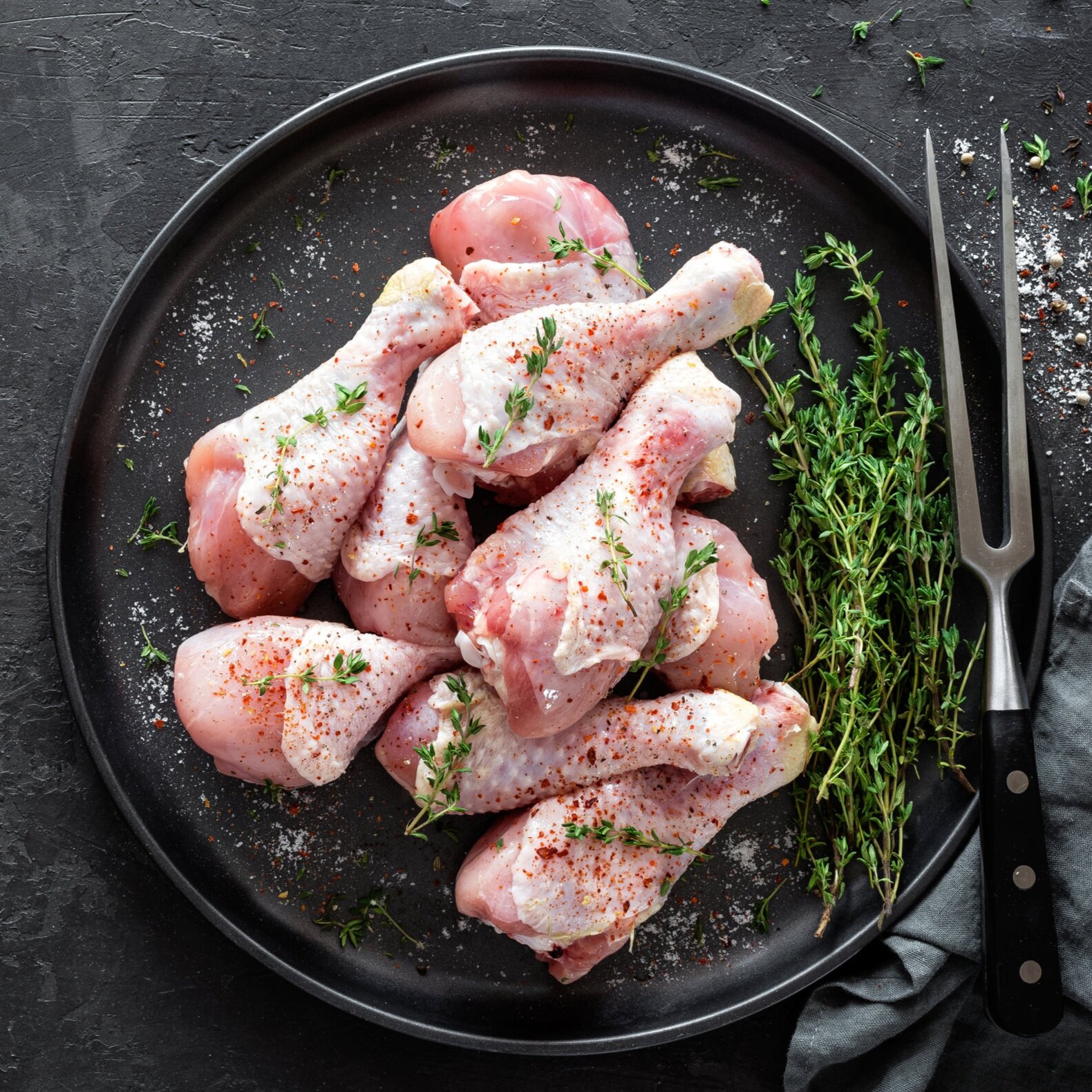 Raw chicken drumsticks seasoned with herbs and spices on a black plate, accompanied by fresh thyme and a carving fork, set against a textured dark background.