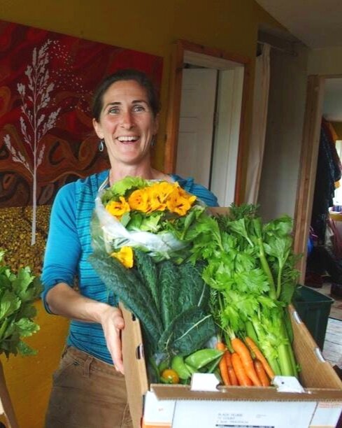 Homer+Hilltop+Alaska+-+carey+with+boxes Smiling woman holding a box of fresh vegetables and flowers, showcasing a vibrant assortment including leafy greens, carrots, and yellow blooms, set against a colorful, artistic background.