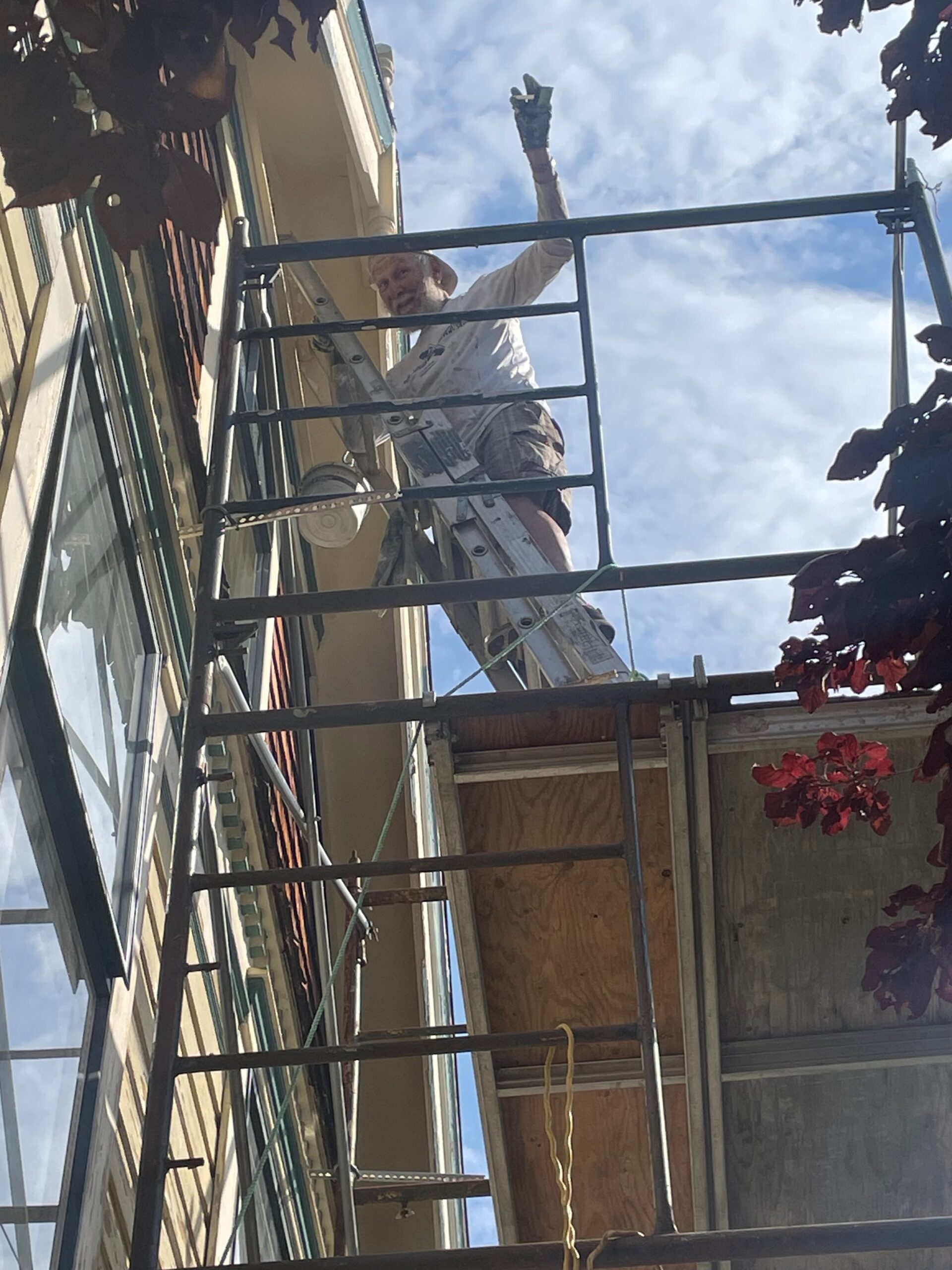 A construction worker on a ladder is painting the exterior of a building while standing on scaffolding. The worker is wearing a hat and gloves, and is gesturing with one hand against a backdrop of blue sky and clouds.