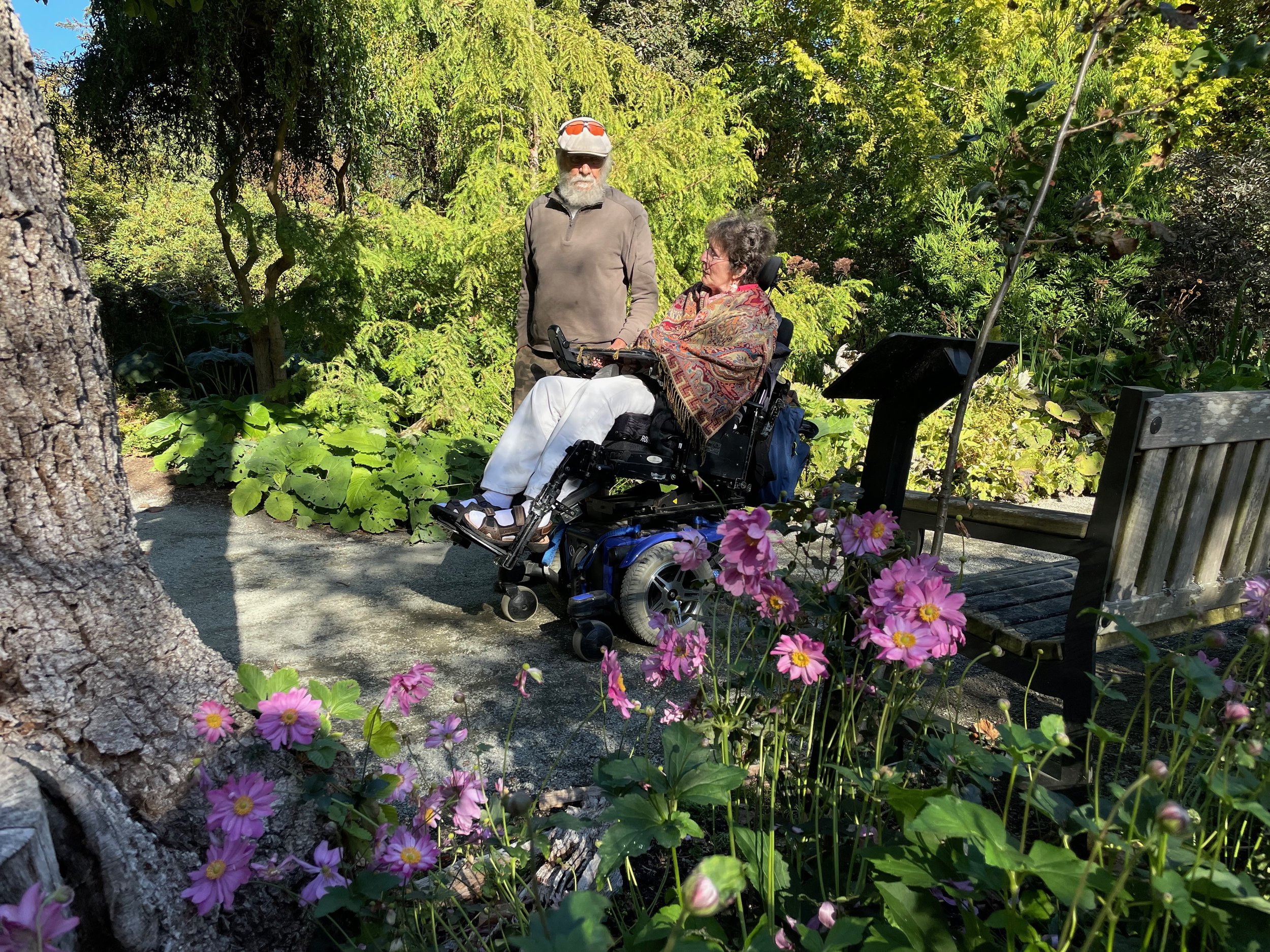 A man standing beside a woman in a wheelchair, both enjoying a vibrant garden filled with colorful flowers and greenery. The scene captures a peaceful moment in a well-maintained outdoor space, highlighting accessibility and companionship in nature.