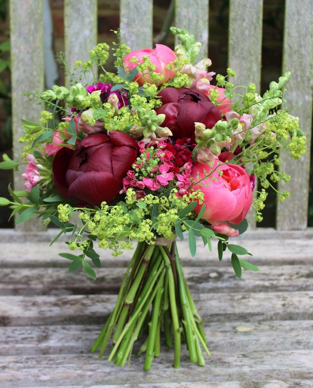 Vibrant floral bouquet featuring deep red and soft pink peonies, accented with snapdragons and lush greenery, arranged on a rustic wooden surface. Perfect for weddings, celebrations, or as a decorative centerpiece.
