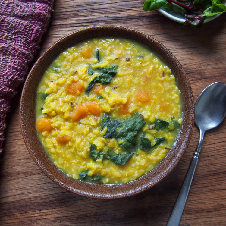 Bowl of creamy lentil soup with spinach and carrots, served on a wooden table next to a spoon, showcasing a nutritious and hearty meal option.