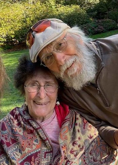 Elderly couple smiling together outdoors, surrounded by greenery, showcasing a joyful moment of companionship. The woman is wearing glasses and a colorful patterned shawl, while the man has a beard and is wearing a cap with sunglasses on his head.